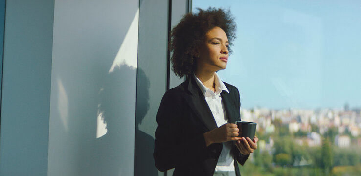 Confident african american businesswoman in a black blazer pauses with coffee by an office window, gazing at the city skyline while planning strategy and future success