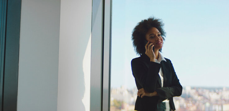 Afro businesswoman making a phone call, standing by large window, having remote business conversation with client while overlooking city view from corporate office - Powered by Adobe