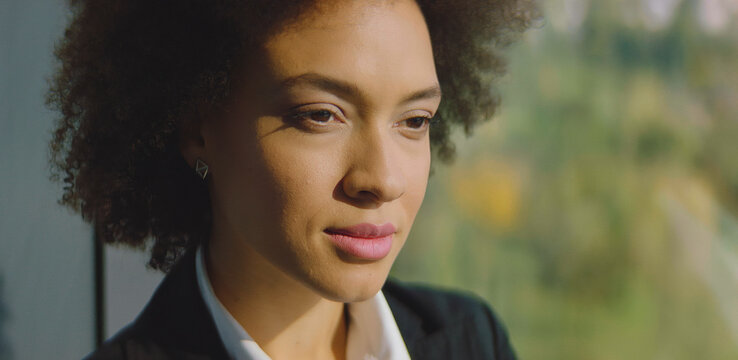 African american businesswoman with curly hair looking thoughtful and focused, standing in a professional setting with natural sunlight highlighting her face, representing success and determination