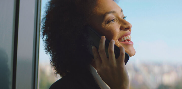 African american woman enjoying a pleasant phone conversation while standing next to a large window in a modern office, illuminated by natural sunlight