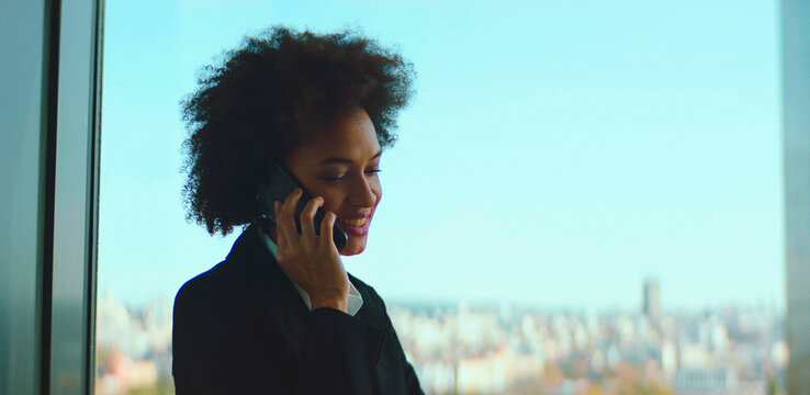 African american businesswoman communicating on her mobile phone, having a pleasant conversation while standing by a large window overlooking a city skyline