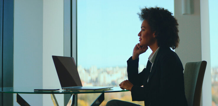 Professional businesswoman sitting at office desk, resting chin on hand and thoughtfully looking out a large window at the city skyline, demonstrating contemplation and future planning - Powered by Adobe