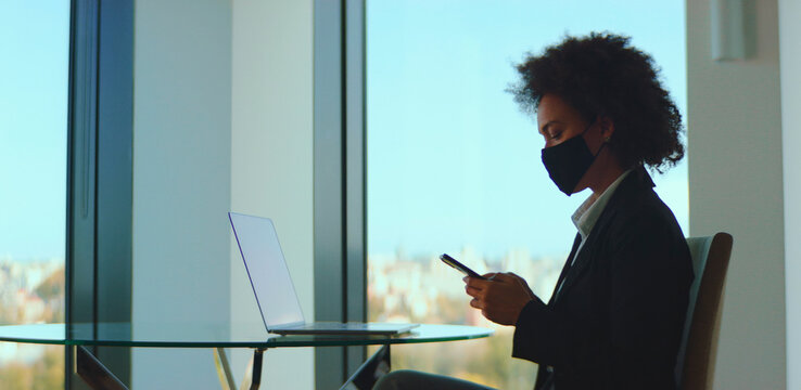 Black businesswoman wearing a protective face mask and formal attire, sitting by a panoramic window, concentrating on her smartphone while a laptop sits opened on a glass table