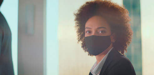 Professional black businesswoman with afro hair wearing a protective face mask while working in a modern corporate office environment during a global health crisis