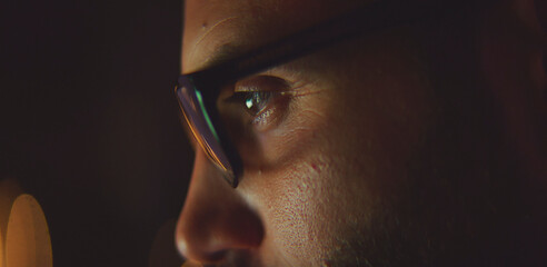 Man with stylish glasses lit by a blue screen glow, intently focusing on data in a dark home office — representing technology, concentration, late-night work and digital engagement