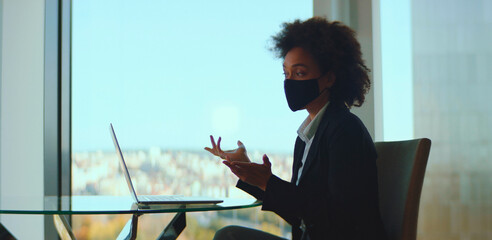 Black businesswoman in a modern office wearing a protective face mask while conducting a laptop video conference, gesturing during a virtual meeting with city view daylight