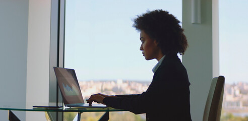 Young businesswoman with afro hair focused on laptop, analyzing financial data from a high-rise office with panoramic city view, embodying productivity and professional ambition