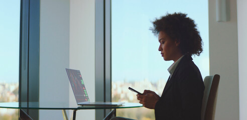 Businesswoman focused on smartphone while analyzing financial charts on a laptop at a glass desk with city view, representing digital trading, investment and market analysis