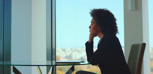 African american businesswoman sitting at a glass table in a modern office, thoughtfully looking out the window at the city skyline while considering professional challenges and career prospects
