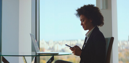 Young professional businesswoman sitting at a glass table near a large window, using a smartphone and smiling, with a laptop displaying financial data beside her and a city view in the background