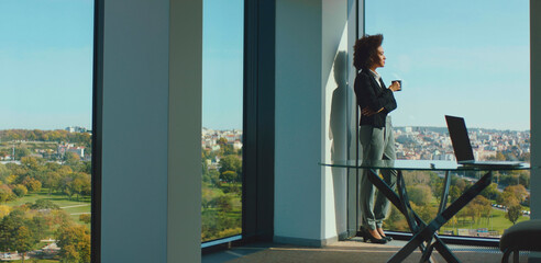 Professional businesswoman standing by the large office window, holding a coffee cup, taking a thoughtful break from work, enjoying a panoramic city and park view on a bright day