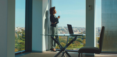 Young businesswoman with curly hair standing by a large office window, holding a coffee cup, looking out at the sprawling city skyline below while taking a thoughtful break from work © Videophilia