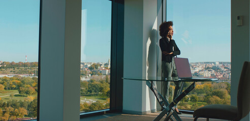 Businesswoman on phone by a large office window, gazing at a green city skyline and park, embodying confident professional communication, leadership and urban workplace vision