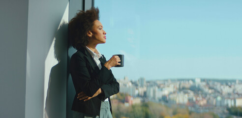 Young professional businesswoman of color standing beside a large office window, holding a coffee...