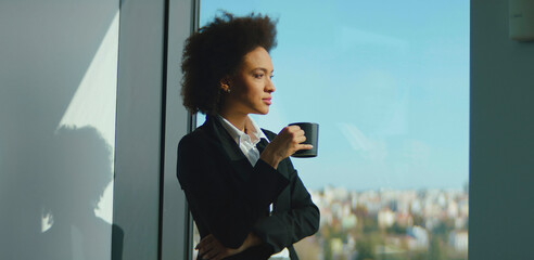 Young black businesswoman standing near a large office window, holding a coffee mug, looking thoughtfully at the cityscape below while taking a moment during a busy workday