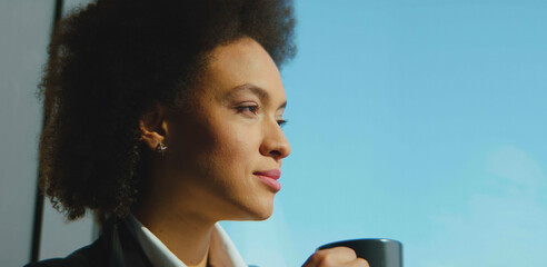 Thoughtful young adult woman with afro hair drinking coffee and looking out a window, enjoying a moment of peace and contemplation under bright natural light