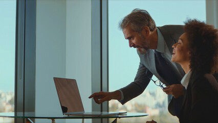 Diverse business professionals collaborate in a bright modern office, experienced man points at laptop while mentoring a smiling young woman, teamwork and knowledge sharing
