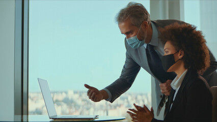 Business colleagues in face masks collaborate over a laptop in a modern high-rise office, discussing strategy and maintaining safety precautions during the covid-19 pandemic