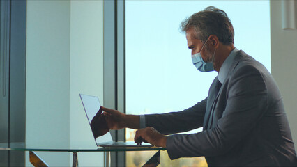Businessman wearing a protective face mask while working diligently on his laptop at a modern office desk, adapting to new normal workplace safety protocols during the global pandemic