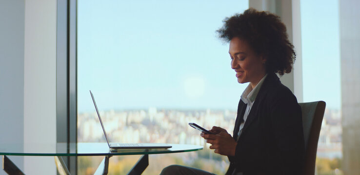 Businesswoman sitting at a glass table, smiling while checking her smartphone with a laptop open next to her, enjoying a bright city view from the office window