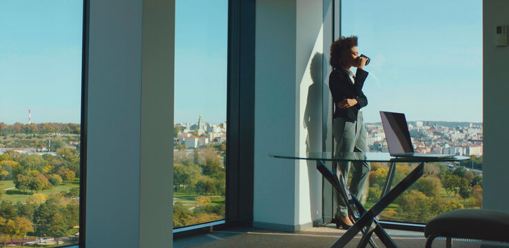 Businesswoman standing by an office window, enjoying a coffee break while observing the expansive city view, a laptop positioned on a modern glass desk nearby, reflecting a moment of contemplation