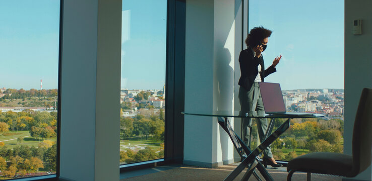 Black businesswoman wearing a face mask standing in a modern office space, talking on a mobile phone with a cityscape and green park visible through large windows - Powered by Adobe