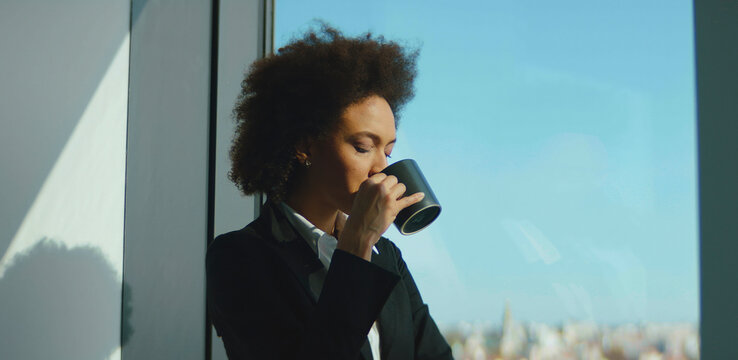 Young businesswoman with afro hair drinking coffee while thoughtfully looking out of a large office window, enjoying a relaxing moment and city view during professional workday - Powered by Adobe