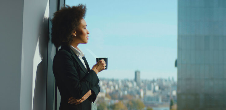 Confident businesswoman with a thoughtful expression standing by a large office window, holding a cup and looking out at the sprawling city, representing ambition and a successful career