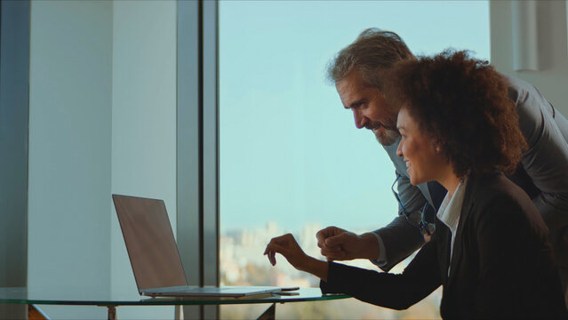 Business colleagues of different generations and ethnicity are collaborating and discussing work together, using a laptop in a bright modern office with a city view