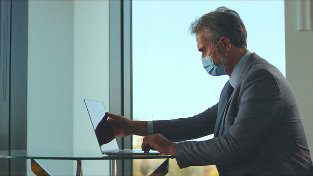 Businessman wearing a protective face mask while working diligently on his laptop at a modern office desk, adapting to new normal workplace safety protocols during the global pandemic - Powered by Adobe