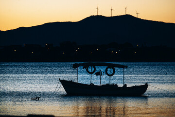 Fishing boat silhouette on calm sea at sunset with wind turbines