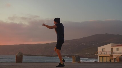 Determined sporty man practicing air boxing overlooking seascape, with mountains in the background and sky at sunset. Fit young man doing interval training in nature