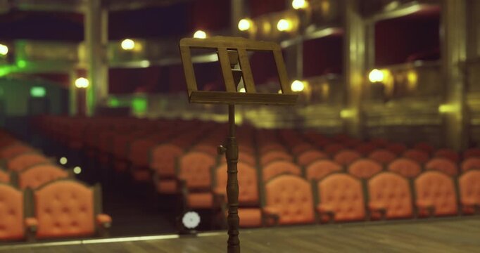 Rows of elegant red velvet chairs fill a grand theater as a solitary music stand awaits on stage.