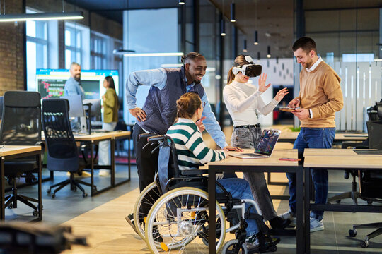 Diverse business team collaborating in a modern office environment. A woman in a wheelchair works on a laptop while colleagues explore virtual reality and digital tools, promoting innovation - Powered by Adobe