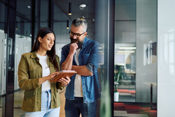 Two business professionals having a focused discussion while reviewing information on a tablet in a bright modern office hallway, symbolizing teamwork, communication, and digital collaboration.