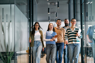 Obraz premium Group of young business team walking together through a modern office hallway, smiling and discussing work. Concept of confidence, teamwork, leadership, and positive workplace culture.