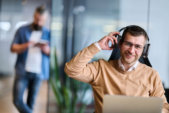 Happy businessman sitting at office desk, wearing headphones and relaxing while listening to music during a break, representing stress relief, balance, and positive work atmosphere in modern office