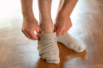 A man putting on a light-colored sock while standing on a floor. Close-up of a man's hands and legs pulling a light gray or beige sock onto his foot, standing on a linoleum floor