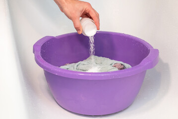 A hand pouring laundry detergent into a basin for hand washing. A hand pouring white laundry detergent or bleach into a purple plastic basin with clothes, care, hygiene, and daily domestic labor