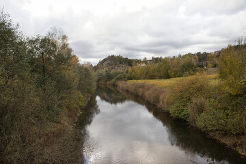 Calm autumn river surrounded by colorful trees under cloudy sky, small red roof house in the distance, Sweden countryside landscape