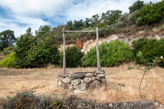 Po&ccedil;o r&uacute;stico de pedra e madeira com um velho c&acirc;ntaro de metal suspenso em meio a um campo seco com vegeta&ccedil;&atilde;o densa sob um c&eacute;u parcialmente nublado
