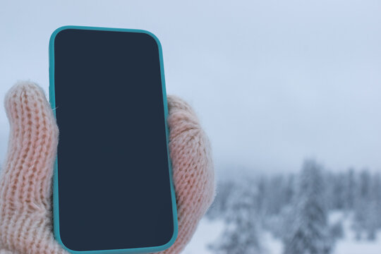 Hand in wool mitten holding smartphone with blank dark blue screen against snowy winter forest. Mockup for design, text, or app presentation in cold season setting