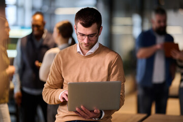 Concentrated male professional using a laptop while standing in a modern open office space, with coworkers collaborating in the background, symbolizing productivity, focus, and teamwork.