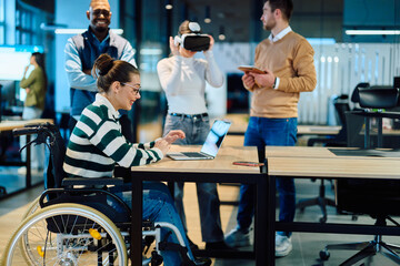 Diverse business team collaborating in a modern office environment. A woman in a wheelchair works on a laptop while colleagues explore virtual reality and digital tools, promoting innovation
