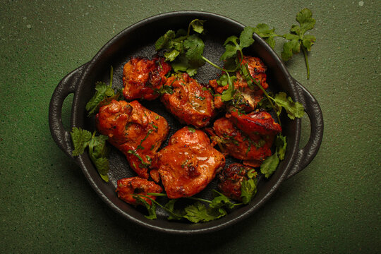 Top view of tandoori chicken, an Indian dish of roasted marinated chicken chunks in spiced curry sauce, served in a cast iron pan on a green background and garnished with fresh coriander leaves