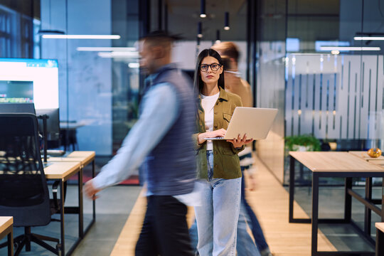 Business woman holding laptop in the middle of a dynamic modern office while colleagues move around, symbolizing focus, leadership, and productivity amid a fast-paced work environment.