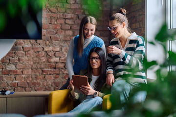 Three cheerful young women sitting in a cozy modern interior, smiling and looking at a tablet screen together, representing friendship, technology, and digital lifestyle.