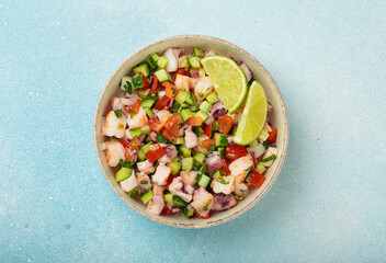 Fresh and vibrant peruvian seafood ceviche with shrimps, octopus, tomatoes, cucumber and parsley, served with lime in a bowl on a light blue background from above, dish of Peru