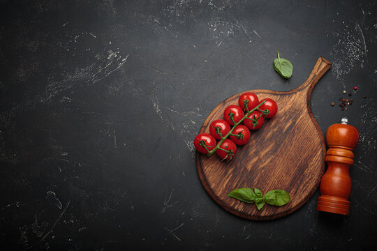 Cherry tomatoes, garlic and fresh basil leaves arranged on a rustic wooden cutting board beside a wooden pepper mill against a dark slate textured background, copy space