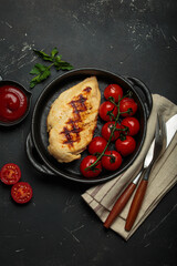 Grilled chicken breast in a cast iron pan on rustic black background, surrounded by roasted cherry tomatoes, ketchup, and cutlery, healthy meal concept